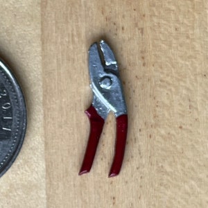 May include: A miniature pair of silver pliers with red handles. The pliers are about the size of a dime.