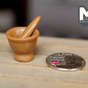 May include: A miniature brown mortar and pestle set beside a silver coin with red detailing. The coin features intricate engravings. The image includes the text "M for MINIATURES".