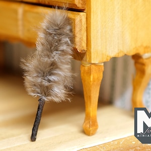 May include: A small wooden chest of drawers with a brown feather duster resting against the front leg. The dresser has a light wood finish and is sitting on a light brown surface.