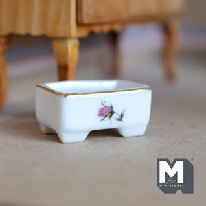 May include: A small white porcelain dish with a gold rim and a pink rose design. The dish has four small feet and is perfect for miniature dollhouse decor.