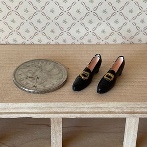 Op de afbeelding: Een paar zwarte poppenhuis schoenen met gouden gespen. De schoenen zijn ongeveer zo groot als een Canadese tien cent munt.