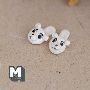 Op de afbeelding: Twee kleine, witte keramische berenhoofden met zwarte en blauwe ogen en lachende gezichten. De berenhoofden hebben afgeronde oren en zijn waarschijnlijk miniatuurbeeldjes of decoratieve elementen. De achtergrond is een getextureerd, neutraal oppervlak.