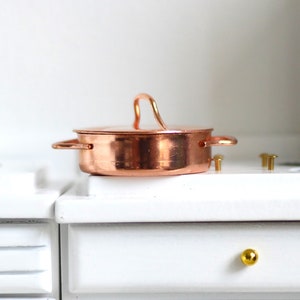 May include: A miniature copper pot with a lid and two side handles, set on a white surface. The pot has a shiny, reflective surface and a curved handle on top. The scene suggests a dollhouse or miniature kitchen setting.
