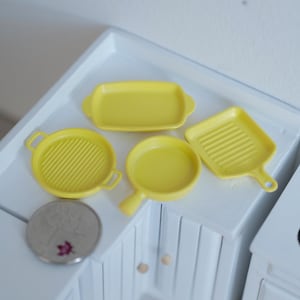 May include: Four yellow miniature kitchen cookware pieces, including a frying pan, a grill pan, a baking dish, and a small saucepan. The cookware is made of plastic and is designed for dollhouse play.