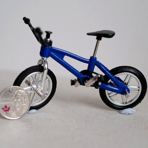 May include: A blue toy BMX bicycle with black tires and a black seat. The bicycle is sitting on a white surface next to a Canadian coin.