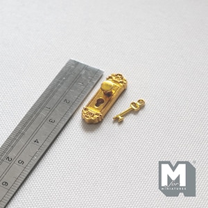 May include: A gold-colored miniature door plate with a knob and keyhole, accompanied by a small matching key. A metal ruler is positioned alongside the items, showing the door plate measures approximately 1.25 inches in total length.