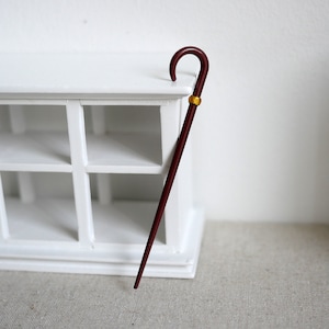 May include: A burgundy walking cane with a curved handle and a gold-colored band. The cane is leaning against a white cabinet with multiple square openings. The cane is a classic accessory.