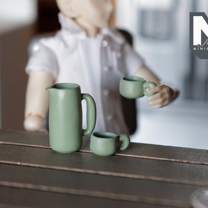 May include: A miniature green ceramic pitcher and two matching mugs sit on a dark wooden surface. One mug is held by a doll figure. The logo "M for MINATURES" is in the upper right corner.