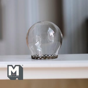 May include: A clear glass lampshade with a decorative metal base. The lampshade is spherical and sits on a white surface. The base has a scalloped edge. The logo "M for MINIATURES" is in the lower left corner.