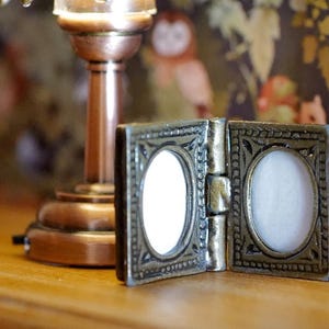 May include: A miniature, open locket in an antique style, with a bronze-toned, decorative frame. The locket features two oval spaces with white interiors. It is hinged and rests on a wooden surface, with a lamp and patterned backdrop.
