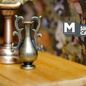 May include: A small, ornate, gold-colored metal vase with a decorative design. The vase is sitting on a wooden surface.