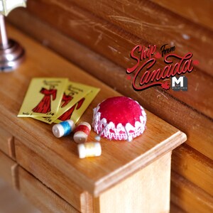 May include: A miniature wooden dresser with three drawers, a red pincushion with white lace trim, spools of thread, and three yellow paper patterns with red dress illustrations. The text "Ship from Canada" is visible on the wooden wall behind the dresser.