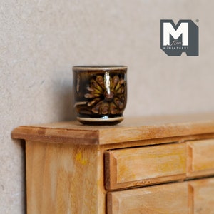 May include: A small brown ceramic pot with a floral design sits on top of a wooden dresser with drawers.