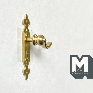 May include: Gold-toned miniature door handle with a decorative design and a keyhole. The handle is attached to a white surface.