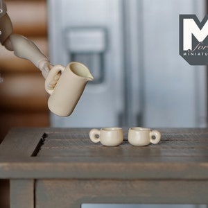 May include: A white ceramic pitcher pouring liquid into two small white ceramic mugs on a wooden table. The pitcher has a handle and a spout. The mugs have handles. The table is brown and has a wood grain pattern. The background is blurred and shows a white refrigerator.