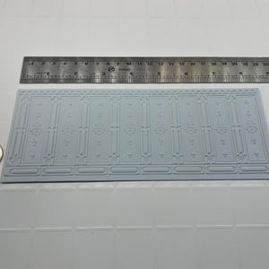 May include: A light grey, rectangular decorative panel with an embossed floral and geometric pattern. A metal ruler is placed above the panel, and a coin is to the left. The panel measures approximately 25 cm in length.