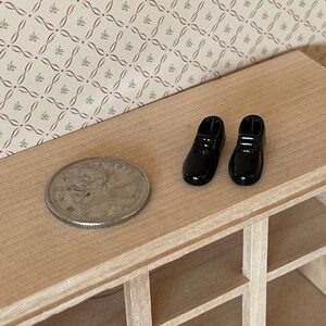 Pu&ograve; includere: Un paio di scarpe in miniatura nere per case di bambole su una superficie di legno con una moneta.