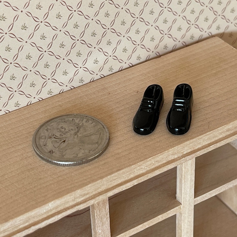 Pu&ograve; includere: Un paio di scarpe in miniatura nere per case di bambole su una superficie di legno con una moneta.