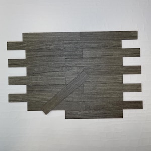 May include: Gray wood-look peel and stick wall tiles arranged in a staggered pattern. The tiles have a realistic wood grain texture and are designed for easy installation. The tiles are rectangular and have a modern aesthetic.
