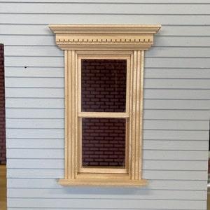 Può includere: Una finestra in legno in miniatura con una cornice di colore chiaro e una parte superiore decorativa. La finestra è inserita in un muro grigio chiaro con pannelli orizzontali. La finestra rivela un interno in muratura.