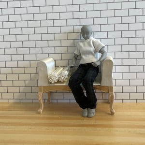 May include: A white doll with black jeans sits on a white bench with a white cushion. The bench has wooden legs and is decorated with a white rolled cushion.
