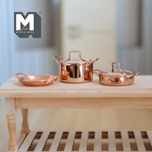 May include: A set of three miniature copper cookware, including a saucepan, a frying pan, and a pot with a lid. The cookware is arranged on a wooden table.