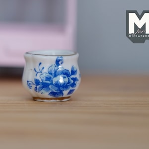以下が含まれることがあります： 青と白の花柄が施された、小さな白い陶器製のプランター。プランターは丸みを帯びた形をしていて、金色の縁取りが施されています。