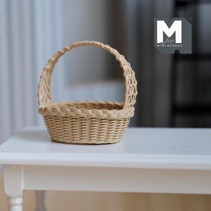Op de afbeelding: Een kleine, beige geweven mand met een handvat. De mand is gemaakt van dunne, verweven stroken, met een gevlochten rand rond de bovenrand. Het handvat is gebogen en ook geweven. De mand staat op een wit oppervlak. Het logo "M for MINIATURES" staat in de rechterbovenhoek.
