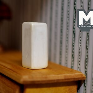 May include: A white ceramic rectangular vase sits on a wooden surface. The vase is smooth and has a slightly textured surface.