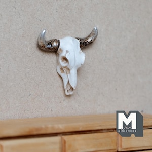 May include: White ceramic bull skull with silver horns mounted on a light brown wall. The skull is a decorative accent for a home or office.
