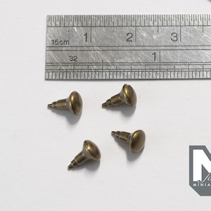 May include: Four small, antique brass-colored metal furniture tacks with rounded heads and pointed shanks, arranged near a metal ruler showing measurements in inches and centimeters to provide a clear sense of their small scale.