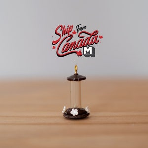 May include: A miniature bird feeder with a brown roof and a clear glass tube. The feeder has a white base with white flowers and two white birds on the sides. The feeder is on a wooden surface. The text "Ship from Canada" is in red lettering above the feeder.