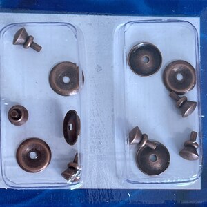 May include: A collection of antique copper-colored furniture hardware, including small knobs and washers, arranged in a clear plastic container. The hardware pieces have a vintage aesthetic, with a slightly tarnished finish.