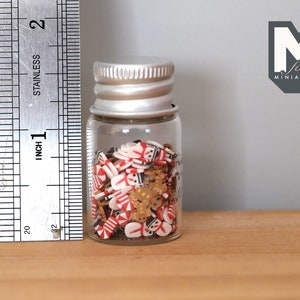 May include: A small glass jar filled with red and white candy cane sprinkles, gingerbread man sprinkles, and gold star sprinkles. The jar is about 2 inches tall and has a silver lid.  The jar is sitting on a wooden surface.