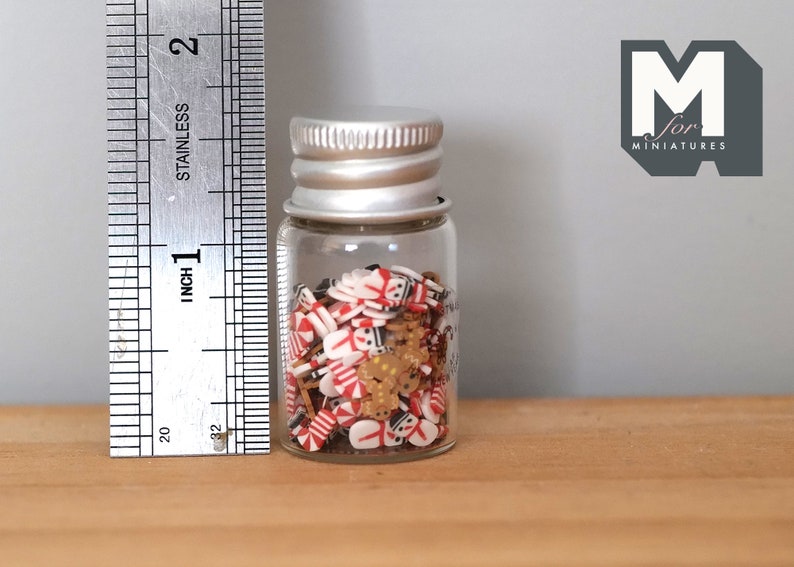 May include: A small glass jar filled with red and white candy cane sprinkles, gingerbread man sprinkles, and gold star sprinkles. The jar is about 2 inches tall and has a silver lid.  The jar is sitting on a wooden surface.