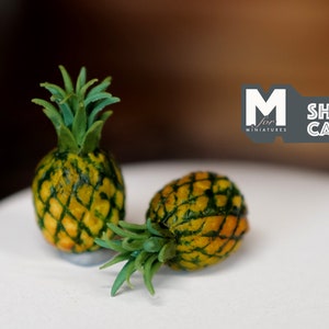 May include: Two miniature pineapples with green leaves on a white surface.  The pineapples are yellow with green markings.  The text "M for Miniatures" and "SHIP from CANADA" is visible in the background.