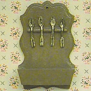 May include: A miniature wooden spoon rack with four small spoons. The rack is a muted gold color and has a decorative top and a small shelf at the bottom. The spoons are also gold and appear to be made of metal. The background is a floral wallpaper.