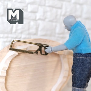 May include: A miniature gold-colored saw with a wooden handle. The saw is being used by a miniature figure to cut a piece of wood.