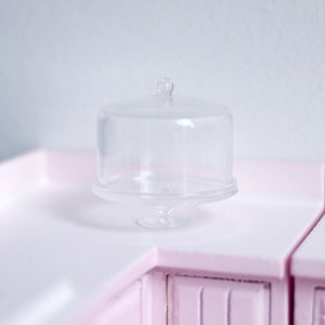 Könnte beinhalten: Ein Kuchenständer aus klarem Glas mit einer Kuppelhaube. Der Ständer hat eine runde Basis und einen Sockel.