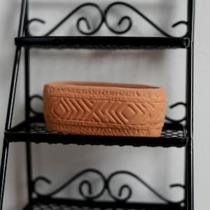 Può includere: Un vaso in terracotta con un motivo decorativo su un ripiano in metallo nero.