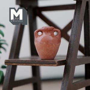 May include: A small, terracotta-colored ceramic pitcher with two handles. The pitcher is sitting on a wooden shelf.