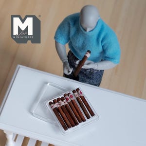 Pode incluir: Uma caixa de plástico transparente contendo vários charutos em miniatura com faixas vermelhas e brancas. Os charutos são marrons com uma ponta de cor mais clara. Uma figura em miniatura com um suéter azul e calças jeans está segurando um charuto. O logotipo "M for MINIATURES" está no canto superior esquerdo.