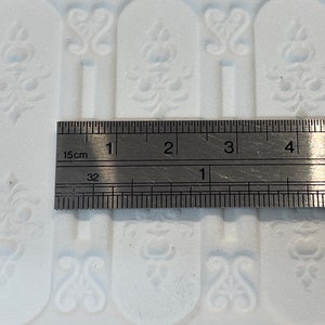 May include: A close-up of a metal ruler measuring 10 cm. The ruler is marked in centimetres and inches. The ruler is placed on top of a white surface with a repeating pattern.