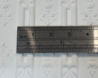 May include: A close-up of a metal ruler measuring 4 inches. The ruler is marked in centimeters and inches. The ruler is placed on top of a white surface with a repeating pattern.