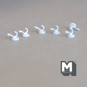 May include: Five small, white, curved hooks are displayed on a neutral surface. Each hook has a rounded base and a curved top, similar to a miniature coat hook. The hooks are evenly spaced. The logo "M for MINATURES" is in the bottom right corner.