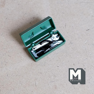 May include: A miniature green toolbox with an open lid, revealing small silver and black tools. The toolbox contains a wrench, pliers, and other tools. The toolbox is on a light brown surface. The logo "M for miniatures" is in the bottom right corner.