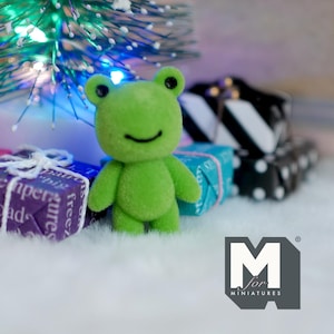 May include: A small, green frog figurine with black eyes and a smiling mouth stands on a white surface. Wrapped gifts in purple, blue, and black and white patterns are in the background, along with a small Christmas tree. The logo "M for MINIATURES" is in the bottom right.