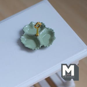 May include: A miniature, three-sectioned serving dish in a soft green hue. The dish has a leaf-like shape with a gold-colored handle in the center. The dish is likely for miniature food items.