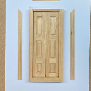 May include: A pair of wooden double doors with a raised panel design. The doors are framed with a simple molding and are set within a larger frame. The doors are light brown in color.