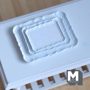 May include: Three white miniature trays with ornate, embossed designs are stacked on a white surface. The trays have scalloped edges and detailed floral patterns. The trays are likely for decorative purposes or dollhouse accessories.